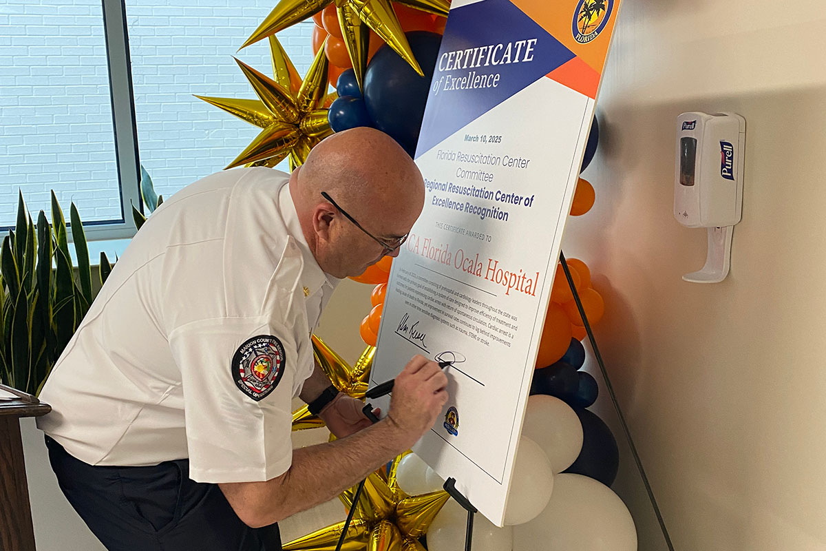 A man in a white shirt leaning down to sign his name on a Certifcate of Excellence poster board